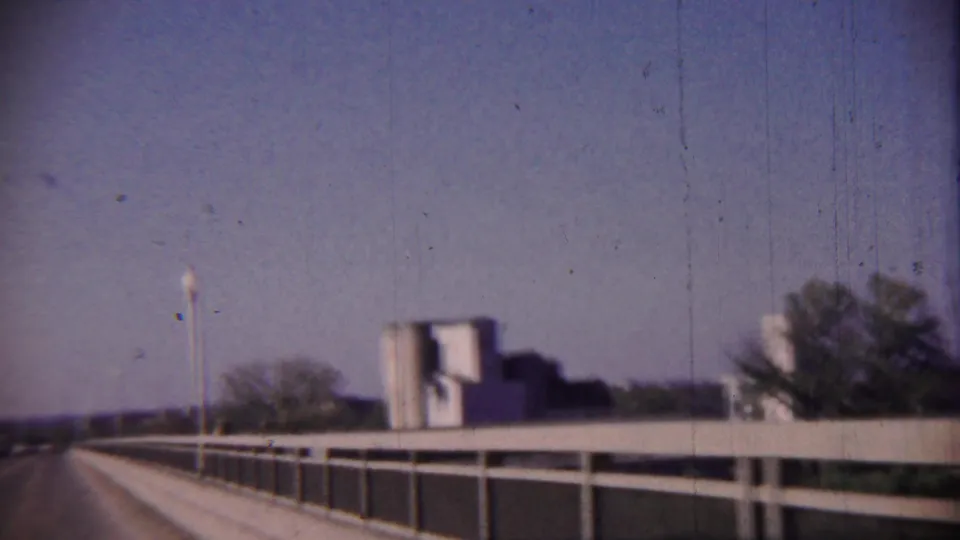 1959 Cheyenne County, Kansas home-movie frame showing a bridge approach with railings, roadside trees, and a distant structure.