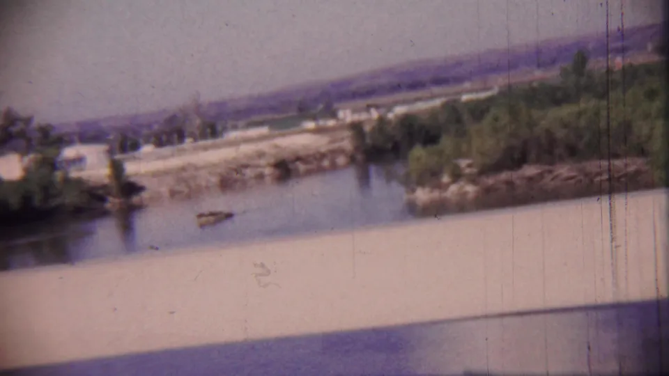 1959 Kansas frame looking from a bridge toward a calm river, wooded banks, distant hills, and a small object on the water.