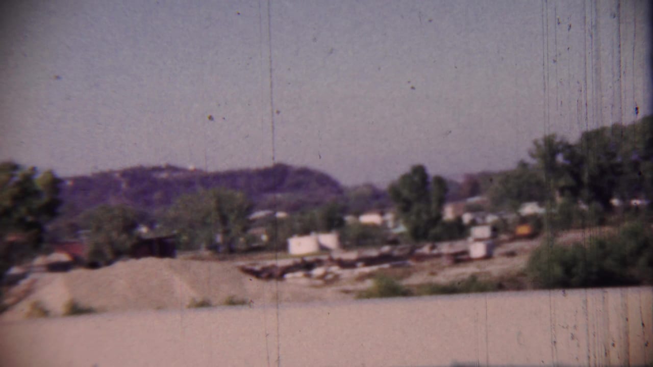 Open gravel area with trees, small structures, and low hills in the distance.