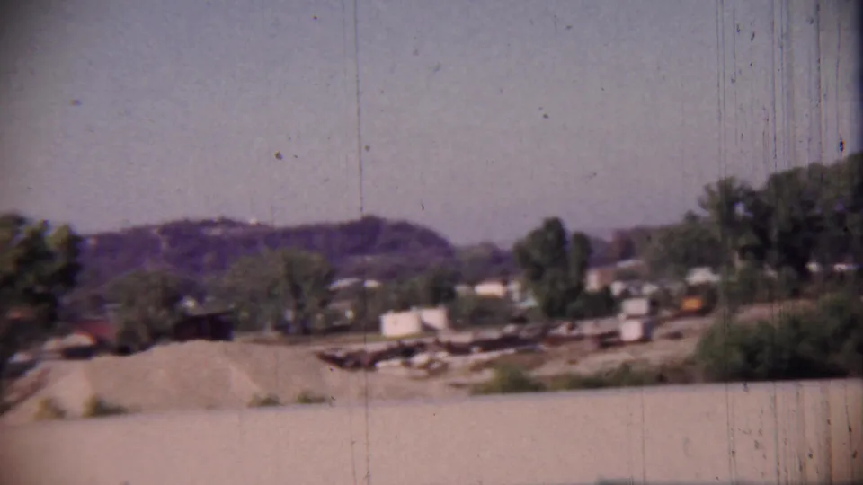 Rural roadside view from the 1959 Cheyenne County, Kansas clip, showing a gravel foreground, scattered buildings, trees, and distant hills.