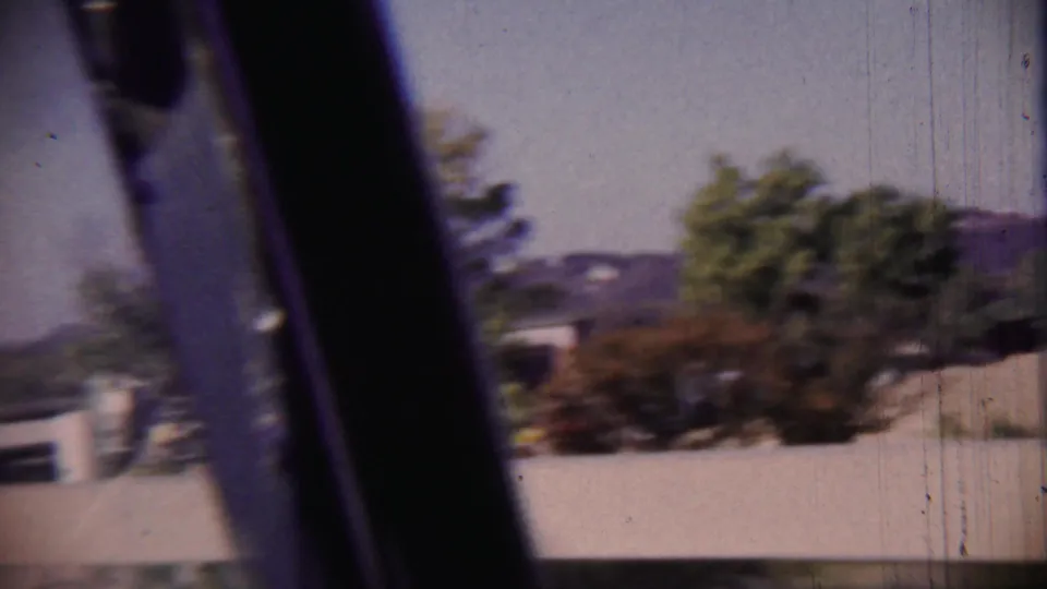 A moving-vehicle frame from the 1959 8mm clip shows a dark window edge, blurred trees, and low roadside buildings.