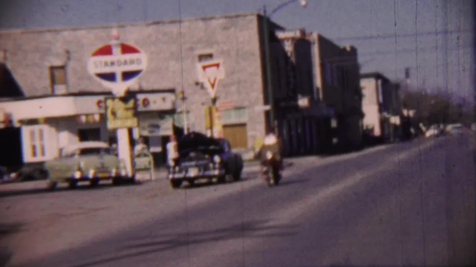 1959 Cheyenne County downtown view with a Standard Oil sign, period cars, a motorcyclist, and low commercial buildings along the street.
