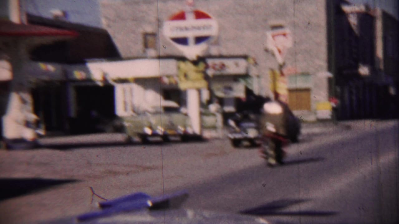 Classic cars and a motorcycle pass storefronts and roadside signs on a sunny street.