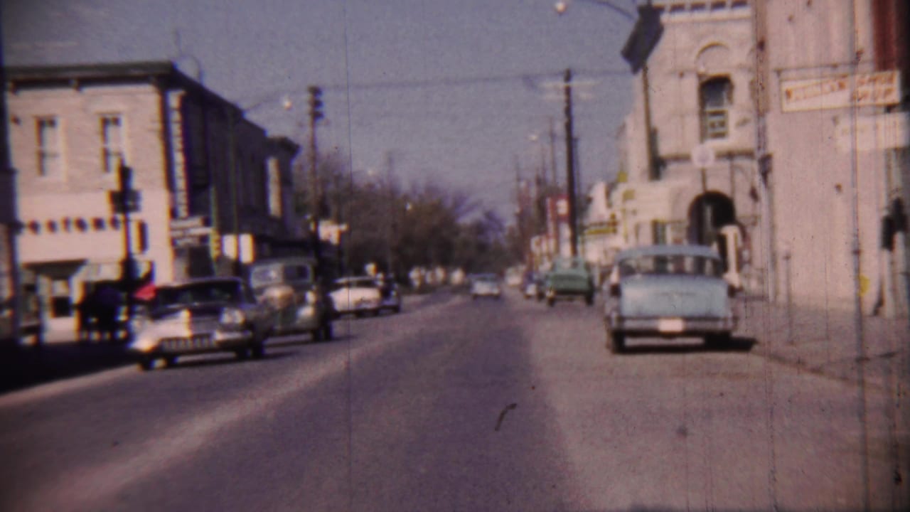 Cars line and move along a commercial street with traffic lights and older buildings.