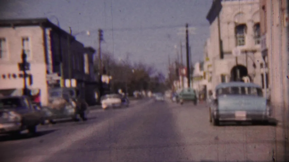 1959 8mm frame showing a pedestrian, classic automobiles, storefronts, and traffic signals in Cheyenne County, Kansas.