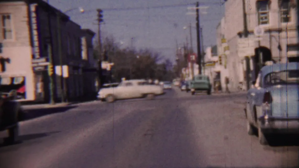End frame of a 1959 Cheyenne County, Kansas street, with a light car, parked vehicles, storefronts, traffic lights, and sidewalks.