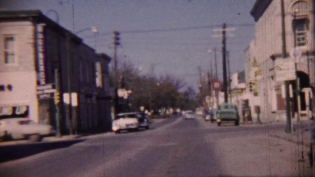 1959 8mm view of a Cheyenne County, Kansas commercial street with parked cars, storefronts, utility poles, and a gently curving road.