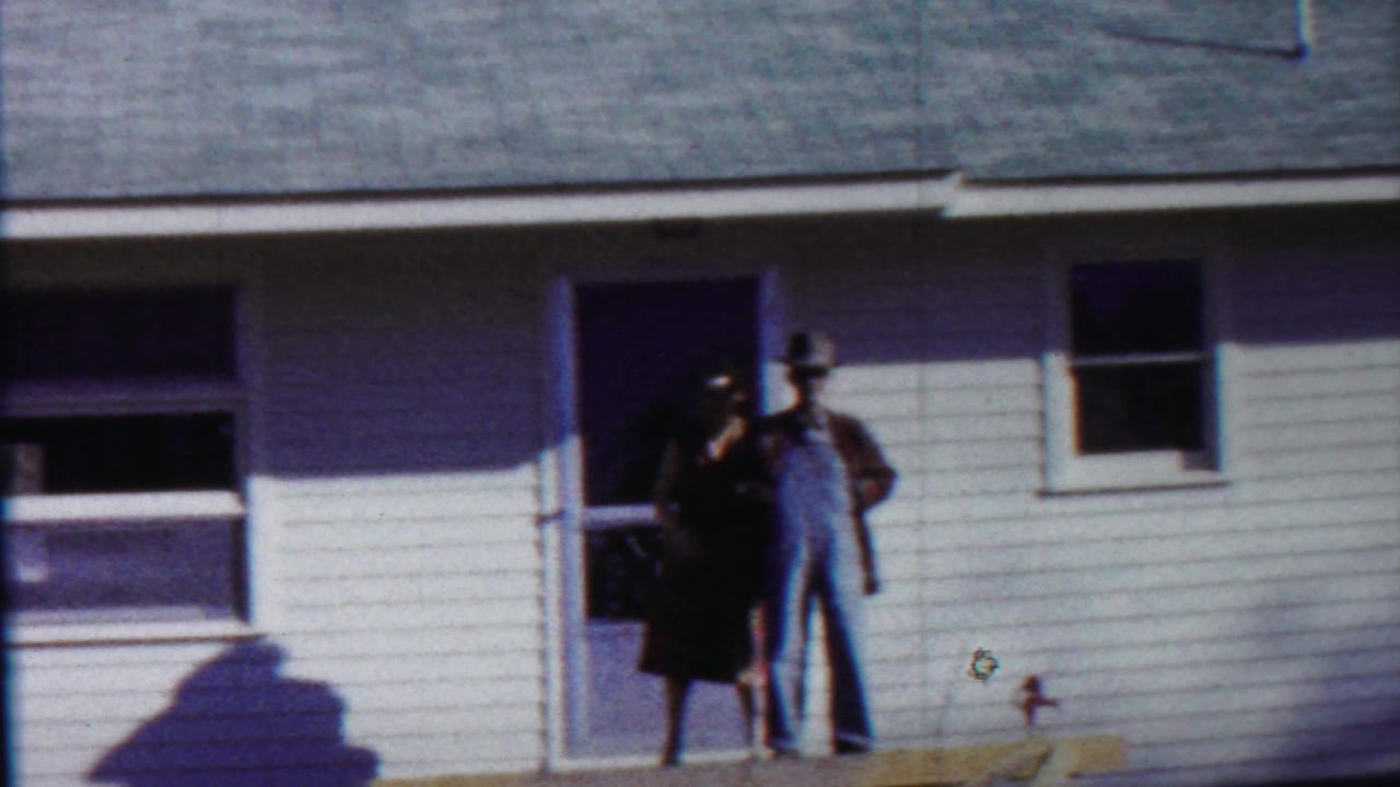 Man in overalls and hat stands near a woman at the doorway of a white-sided house.