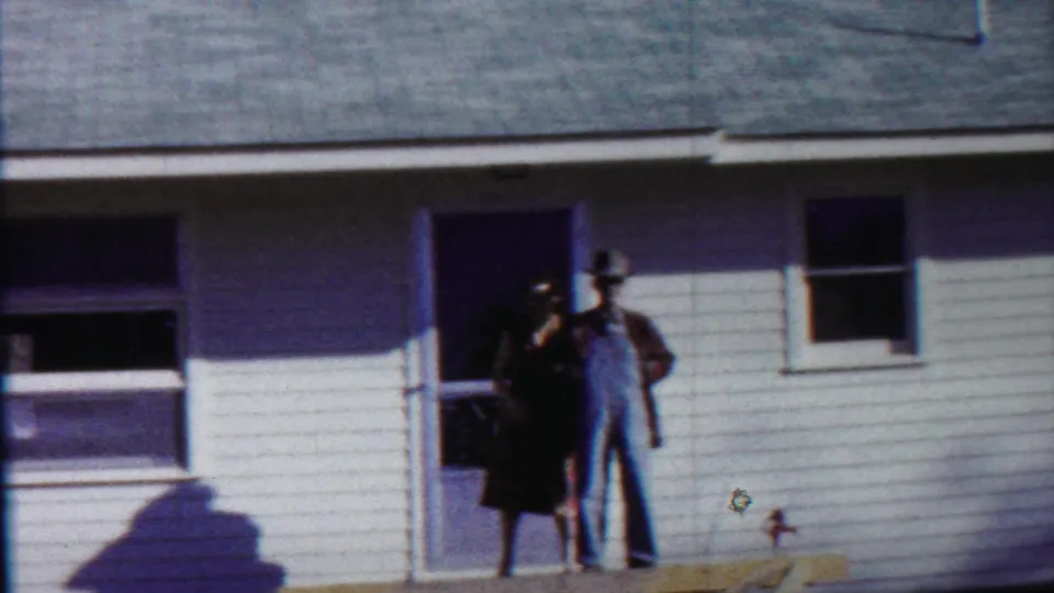 In Saint Francis, Kansas, 1959, a man in overalls and hat stands by a woman at the porch doorway of a white-sided house.