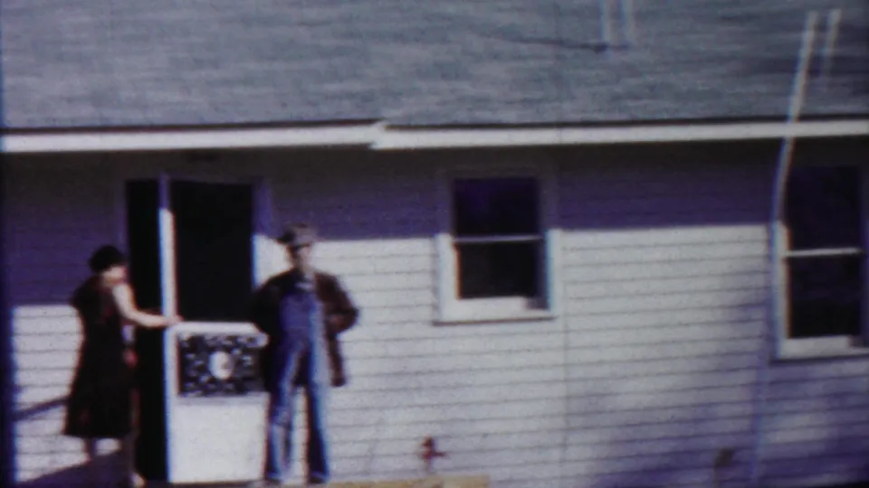 The woman reaches toward the doorway as the man in overalls stands nearby outside the white-sided Kansas house.