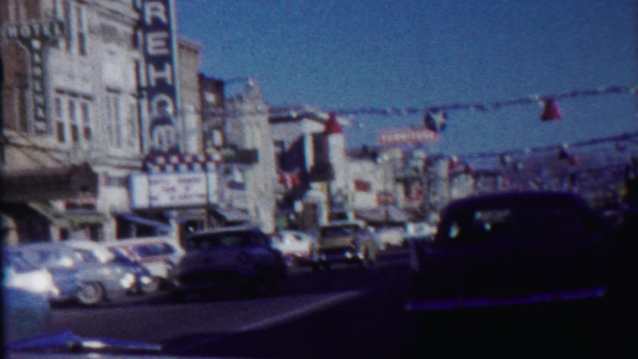 Traffic passes beneath red and blue streamers on a sunlit commercial street.