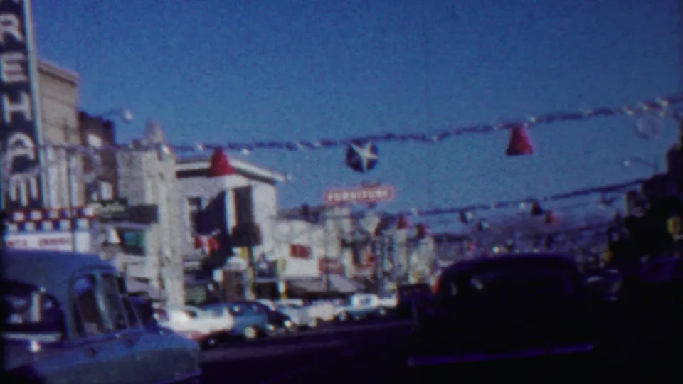 Street-level 1959 view of Saint Francis with classic automobiles, theater marquee edge, storefronts, and red and blue overhead streamers.
