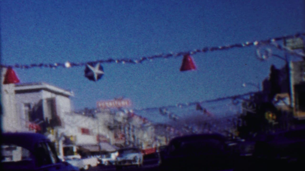 Decorated small-town street with overhead streamers, star ornament, storefronts, and cars under a blue sky.