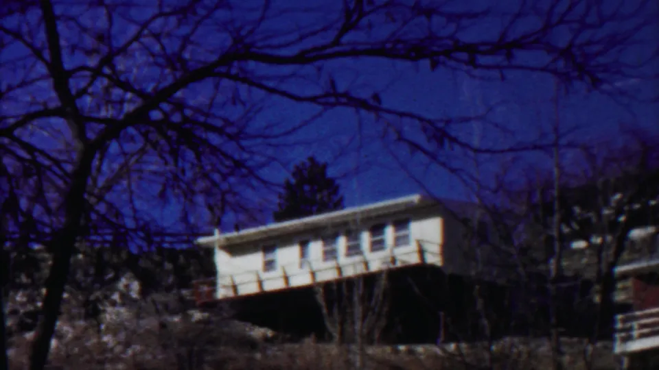 Final 1959 8mm frame of a Saint Francis hillside house, partly framed by tree limbs, brush, and a neighboring roofline.