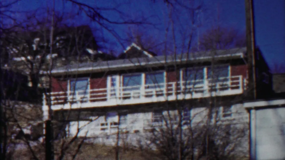 The pan reveals more of the residence, including red wall panels, pale balcony railings, lower stonework, and bare trees.