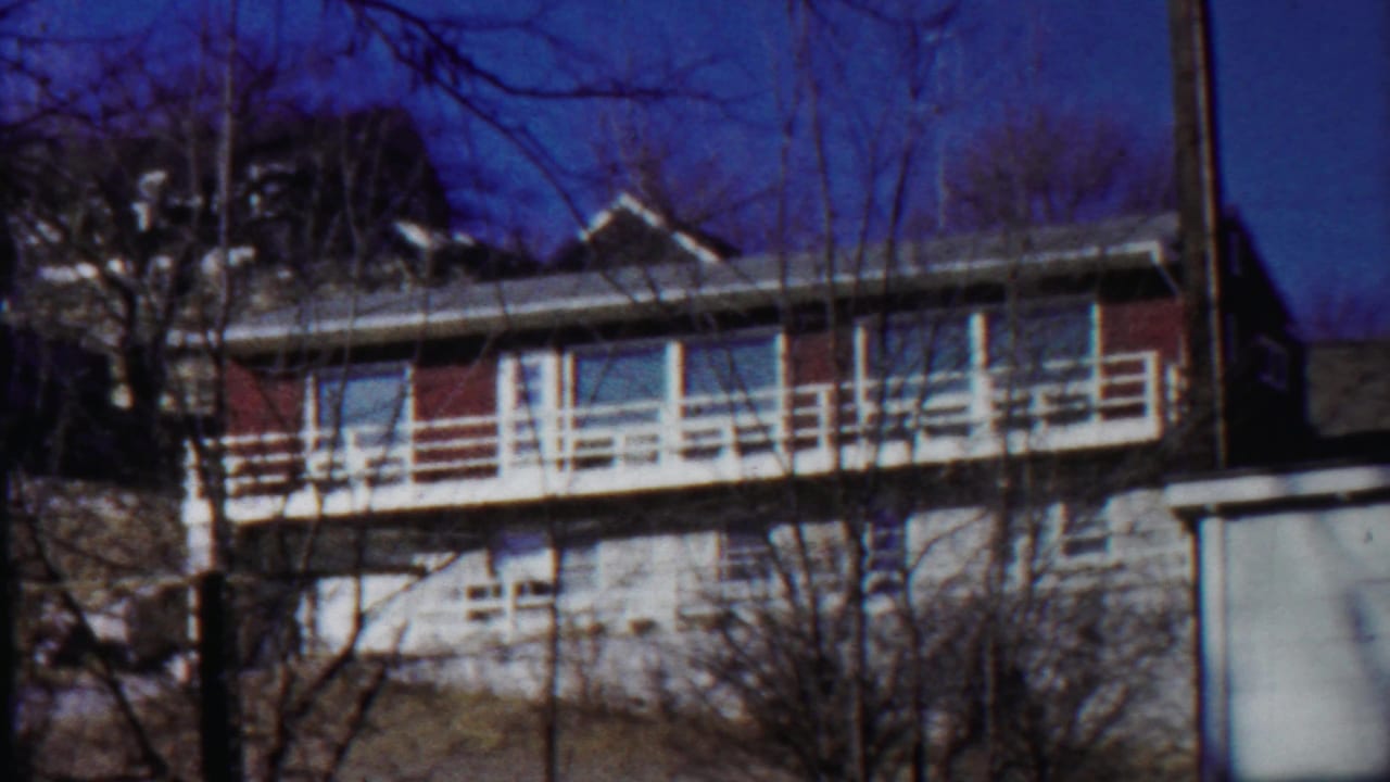 Apartment building exterior with long white balcony, windows, bare branches, and bright sky.