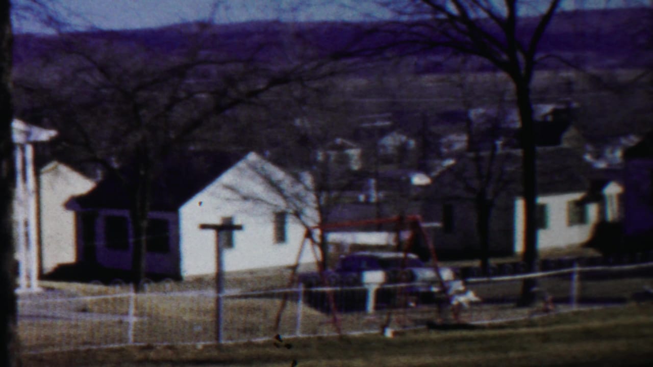 1959 Saint Francis, Kansas home-movie frame showing small white houses, a fenced yard, bare trees, a swing set, and a parked car.