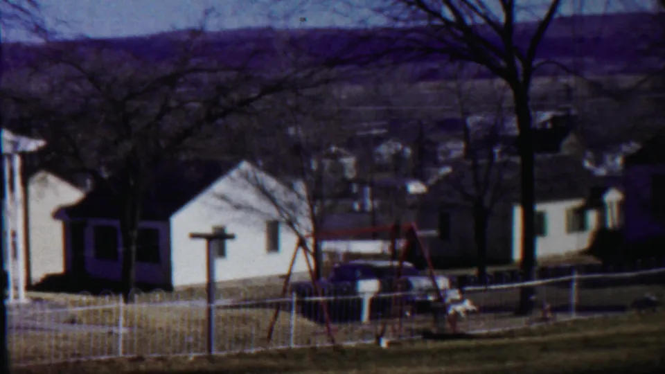 Saint Francis, Kansas residential view with white houses, leafless trees, a white fence, a red swing set, and a parked car in bright sun.