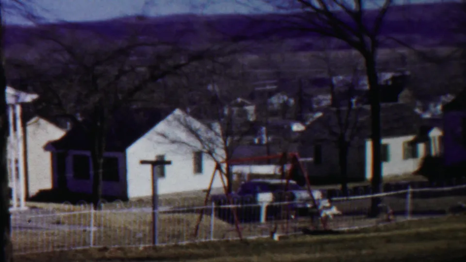 1959 Kansas home-movie still of a sloped neighborhood yard with white houses, fencing, leafless trees, a swing set, and parked car.