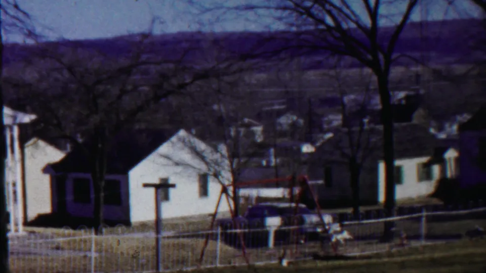 Saint Francis, Kansas frame showing small white houses and fenced yards with bare trees, a red swing set, parked car, and distant hills.