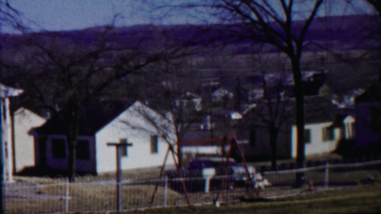 1959 Saint Francis, Kansas home-movie frame showing small white houses, a fenced yard, bare trees, and a red swing set.