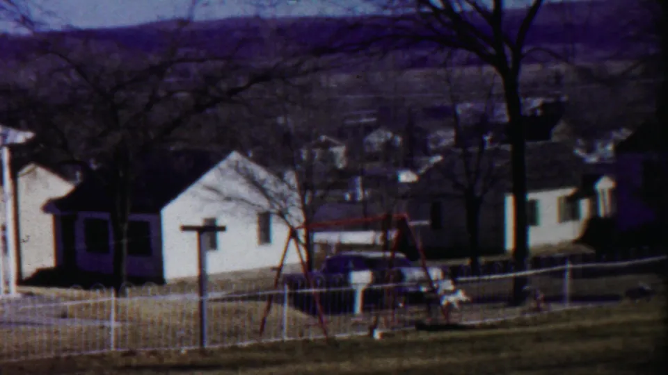 1959 home-movie view of Saint Francis houses with a white fence, leafless trees, a red swing set, and low hills beyond.