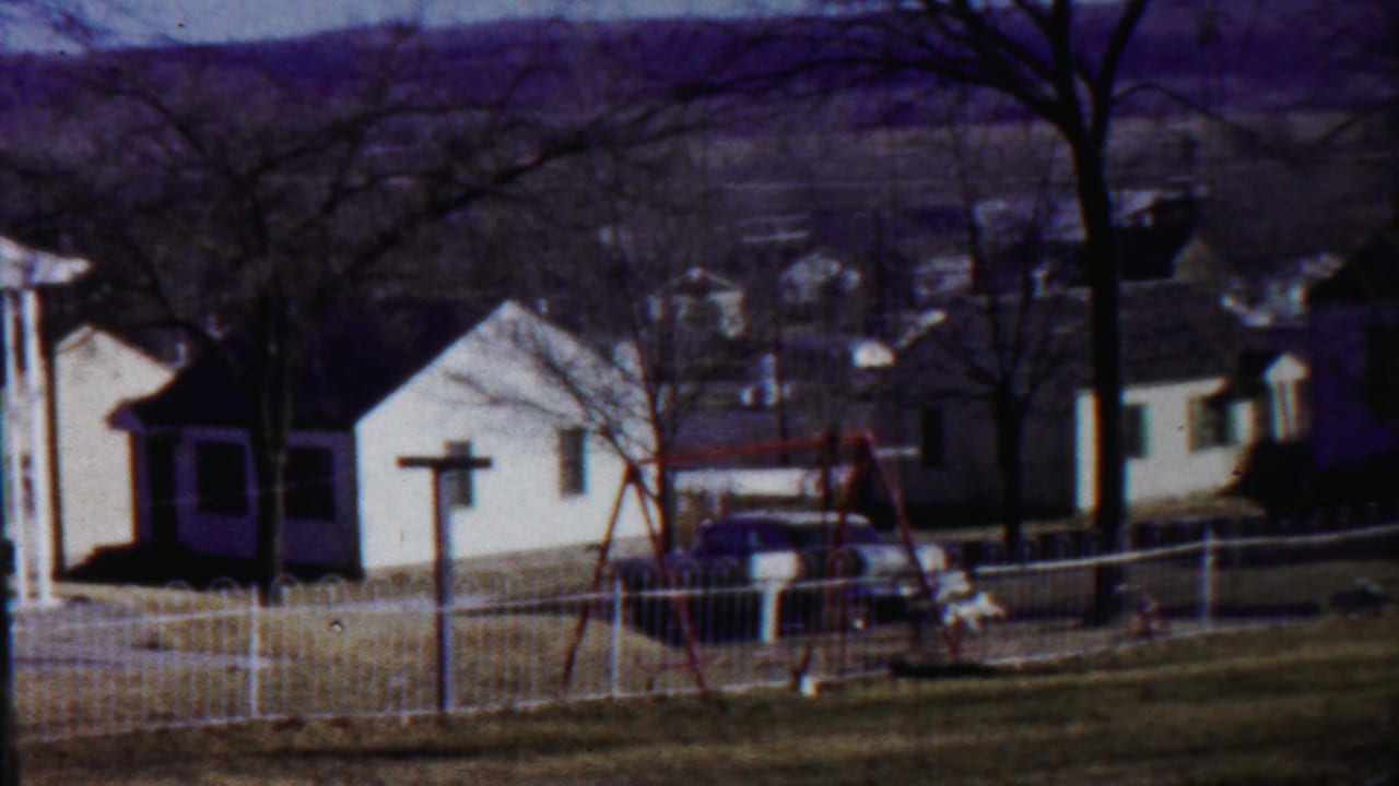 White houses, picket fence, swing set, bare trees, and distant hills beyond a residential street.
