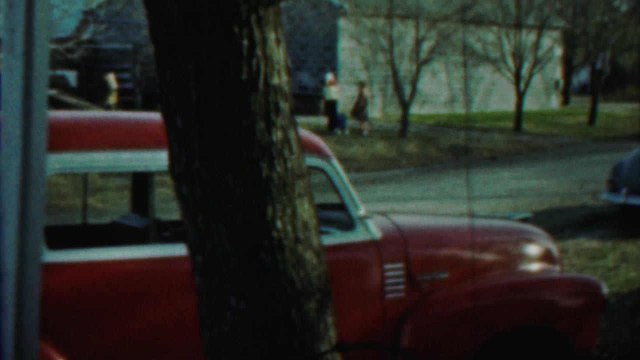 Red vintage car partly hidden by a tree, with a street, lawn, houses, and distant pedestrians.