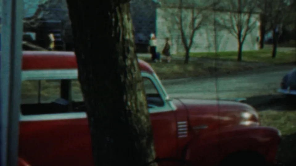 Red mid-century car partly obscured by a tree on a Saint Francis street, with houses, lawn, and distant pedestrians nearby.