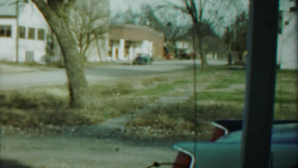 Saint Francis residential street view with a white house, bare trees, grass verge, sidewalk, pole, and a blue car partly visible.