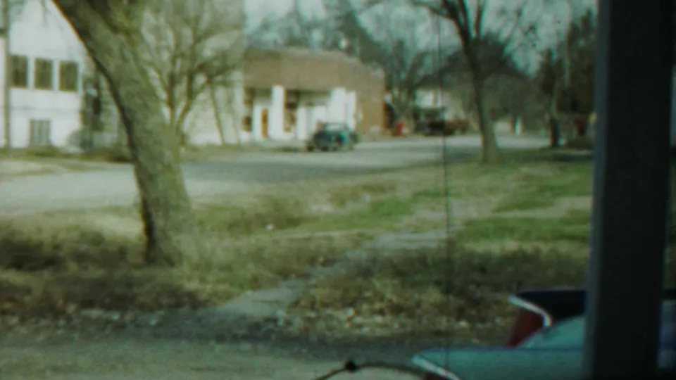 1959 Saint Francis street scene with a white house across the road, bare trees, sidewalk, grass, and a partial blue car foreground.