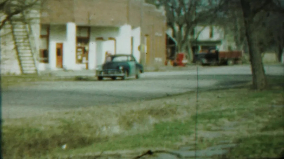 Blue sedan on a Saint Francis street near a brick building with white columns, with bare trees and another vehicle in the background.