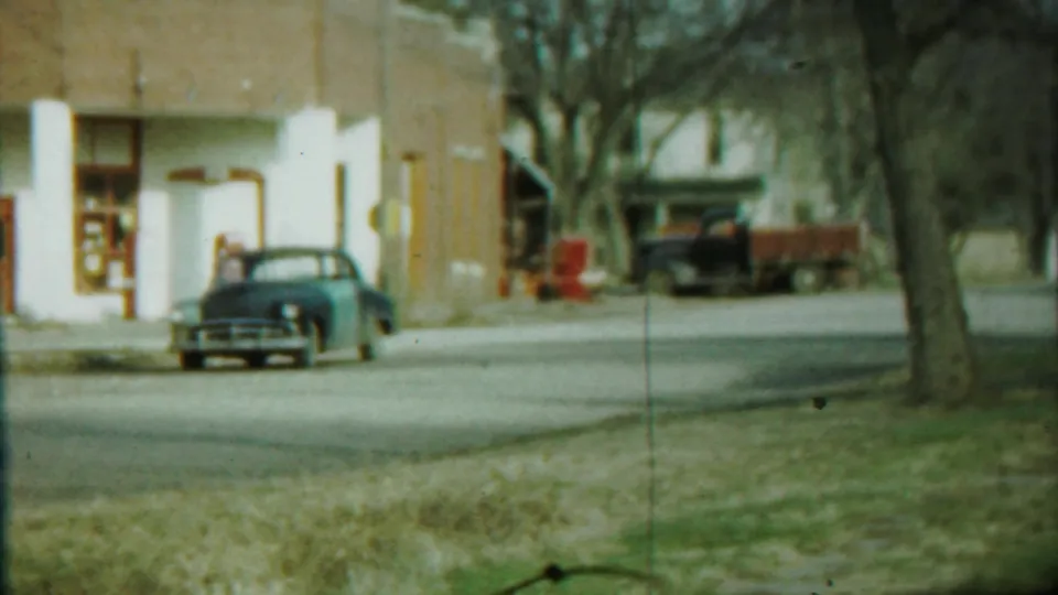 Closer 8mm view of the blue sedan beside the columned brick building, with a darker vehicle or trailer partly visible beyond trees.