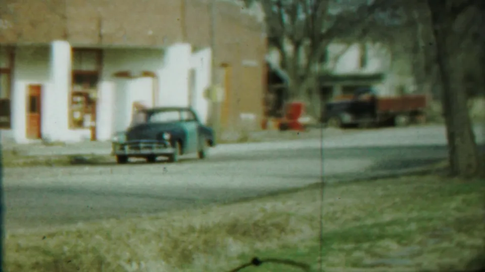Blue sedan centered near a brick building in Saint Francis, Kansas, with grass in the foreground and bare trees along the street.