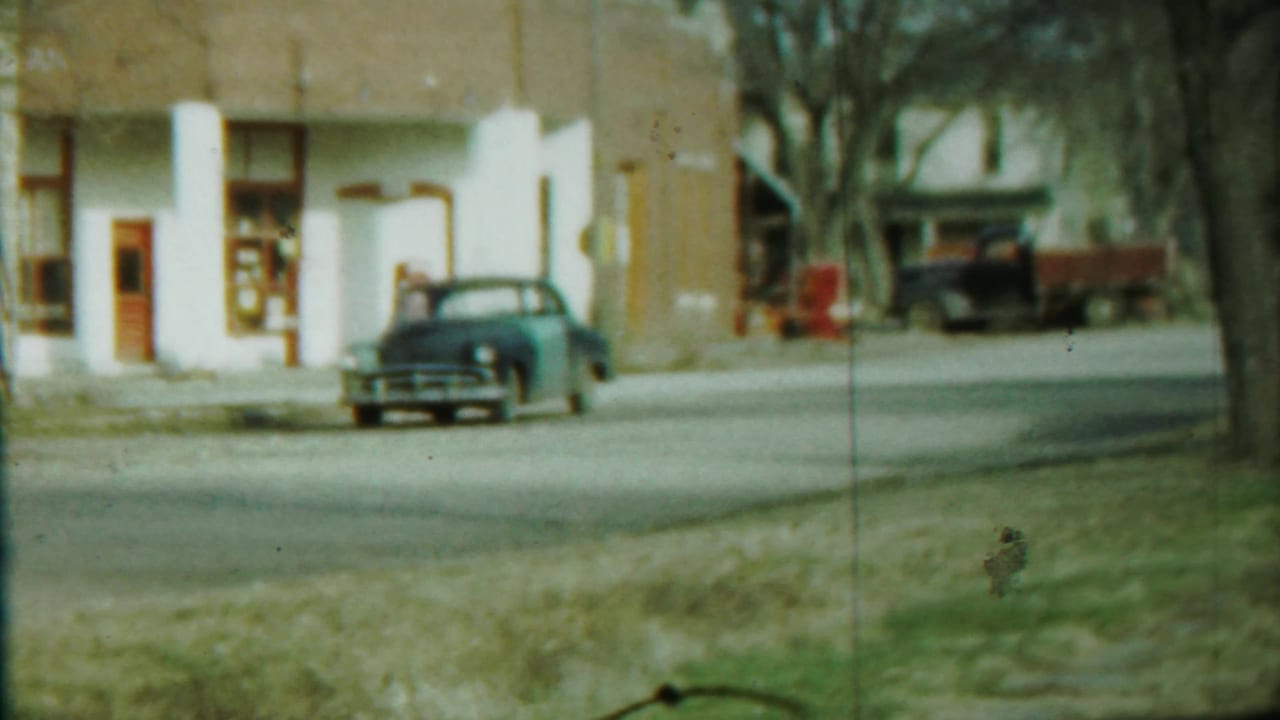 Street view with blue car, white and brown building, trees, and a parked truck in the background.