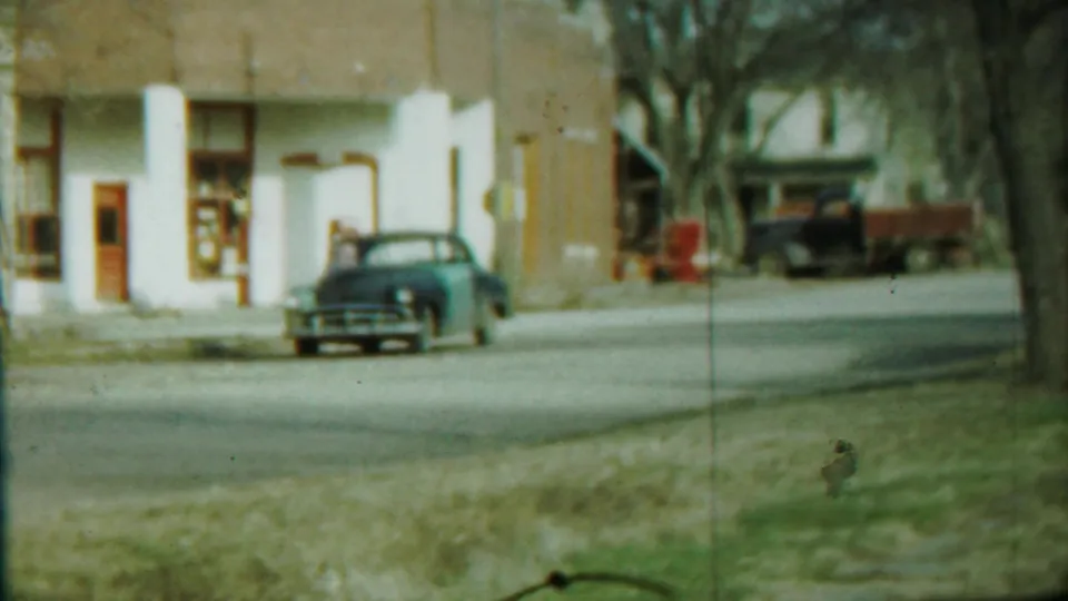 Saint Francis street view with a blue car near a white and brown building and a darker truck farther back, filmed on 8mm in 1959.