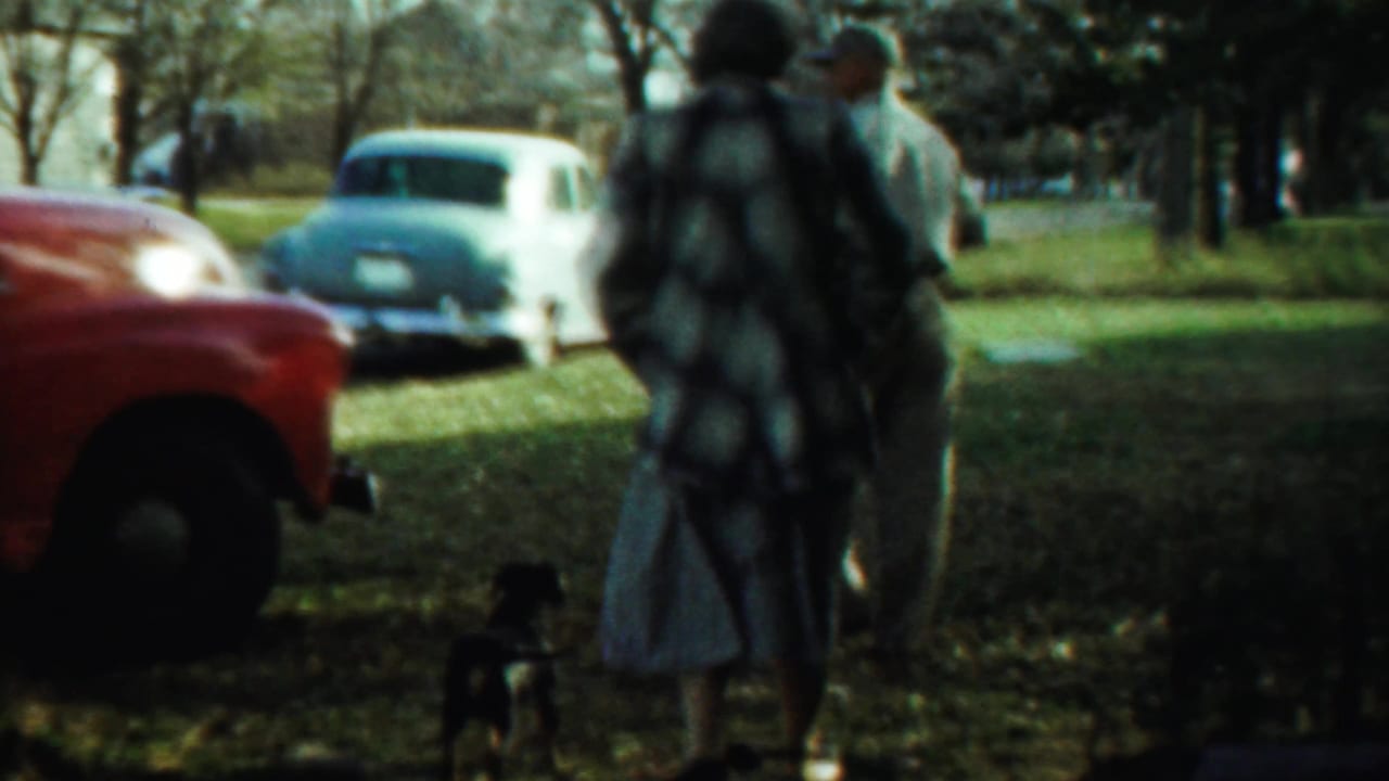 In Saint Francis, Kansas, a 1959 home-movie frame shows adults and a small black dog near a red truck and parked classic cars.