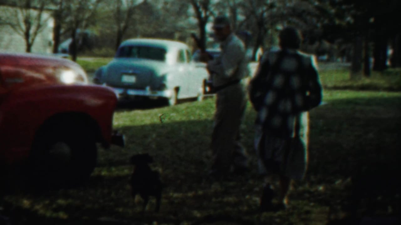 Man holding a rifle stands near a woman, a small dog, a red truck, and parked cars.