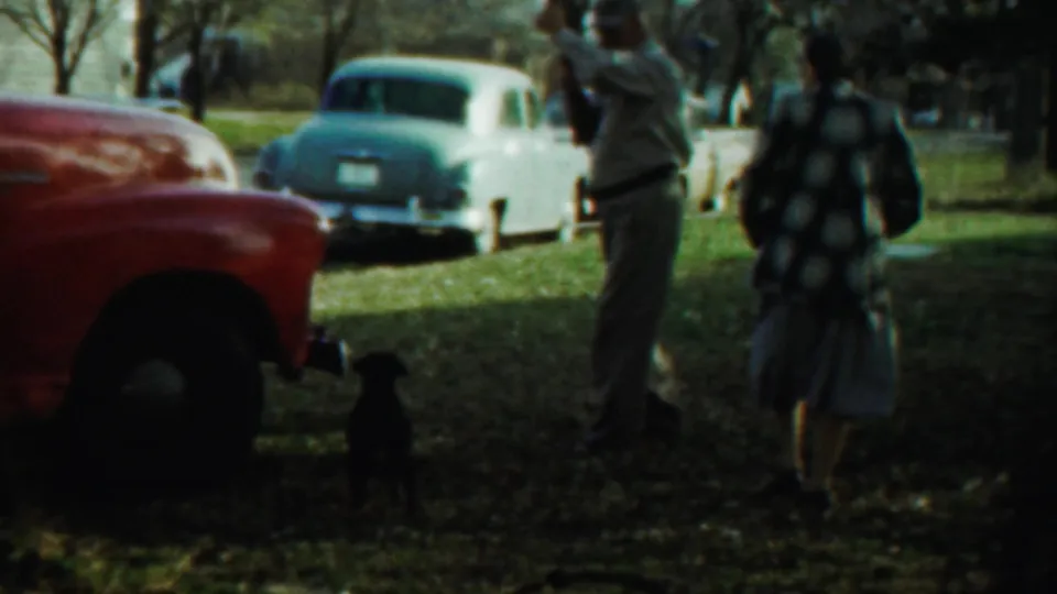 The 1959 Kansas home movie shows a man raising a rifle in a grassy yard as a woman and small black dog stand nearby.