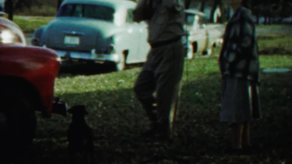 A late-1950s Kansas yard scene shows adults near a small dog, with a red truck and pale classic cars parked behind them.