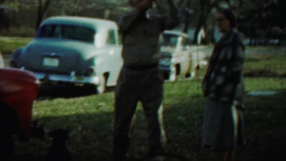 In Saint Francis, Kansas, a man raises a camera while a woman stands beside him near parked 1950s cars on grass.