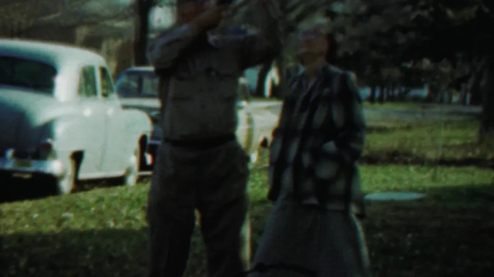 The man keeps the camera raised as the woman stands nearby, with a light sedan and trees visible behind them in 1959 Kansas footage.