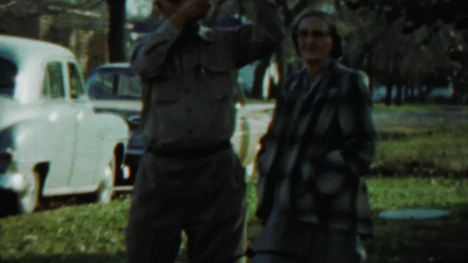 A woman in a plaid jacket and skirt stands beside a man using a camera, with vintage cars and trees in the background.