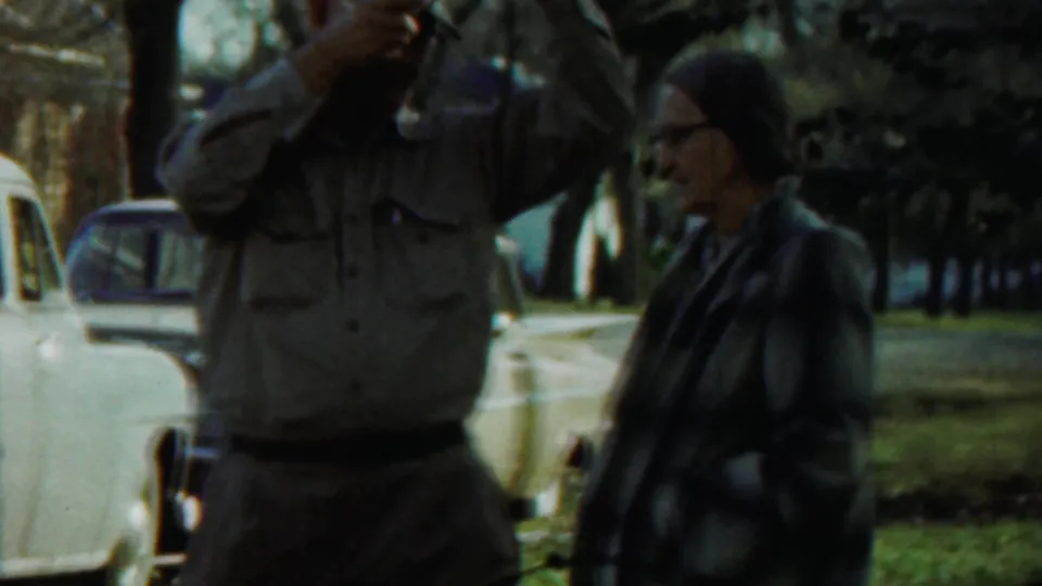 A closer 8mm frame shows the man still using his camera while the woman in plaid stands beside him near parked mid-century cars.