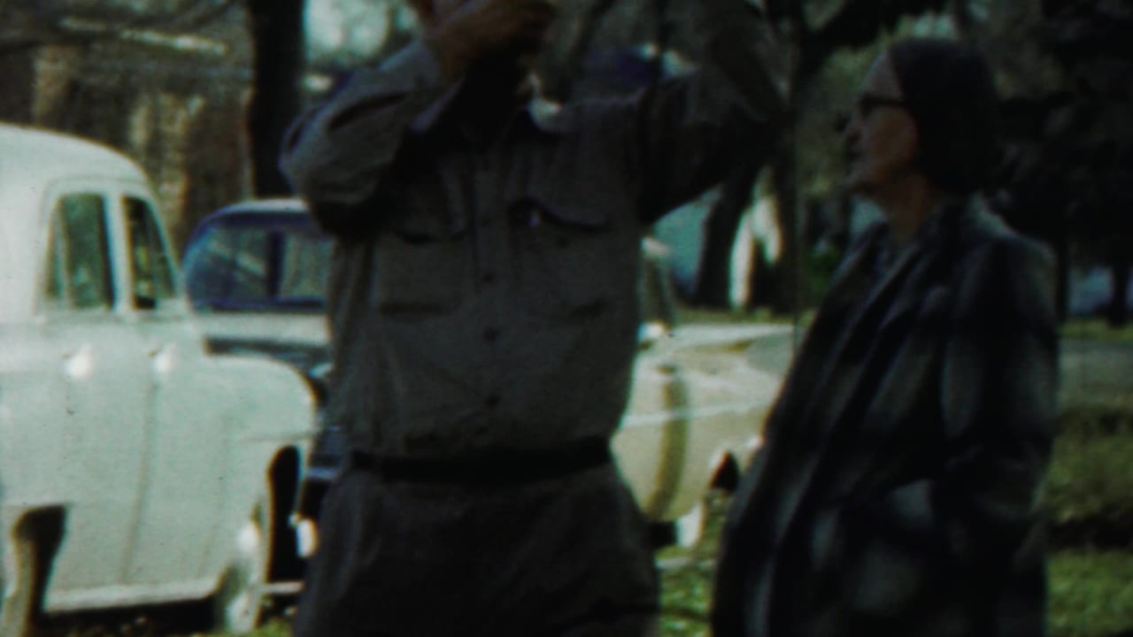 Uniformed man holding a long object near a woman and parked cars in a grassy park.