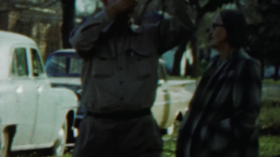 In Saint Francis, Kansas in 1959, a uniformed man stands near a woman in glasses beside parked mid-century cars in a park.