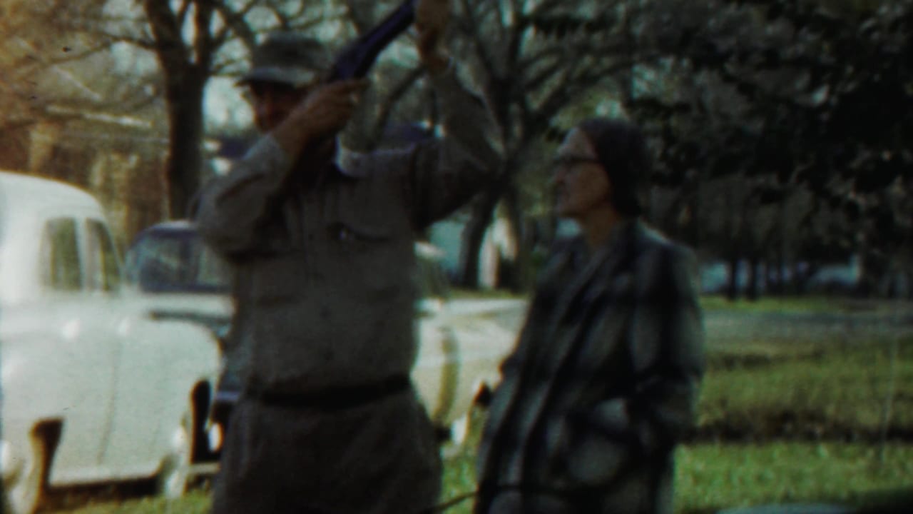 Uniformed man raises a long object while a woman watches in a park with cars behind them.