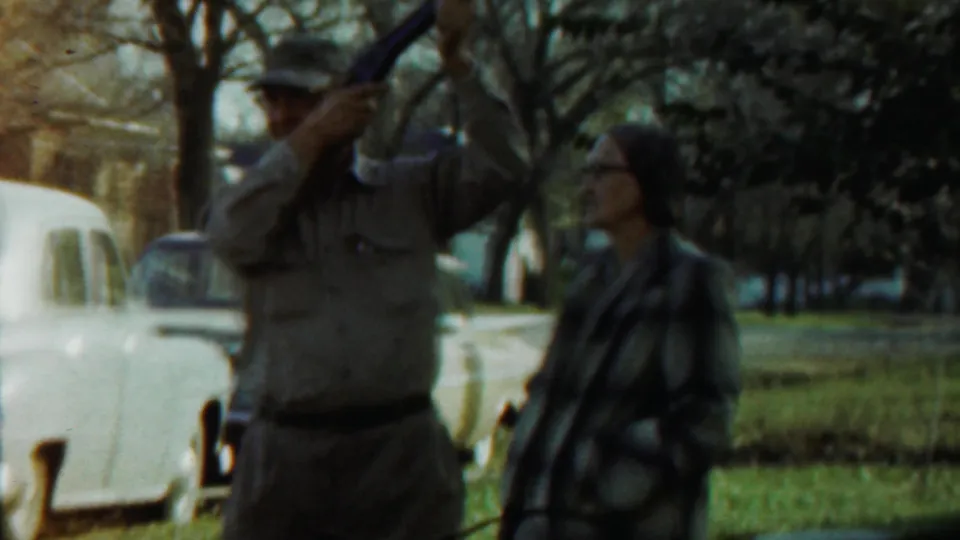 A likely police officer raises a long training object as a woman stands nearby in a 1959 Saint Francis park scene.