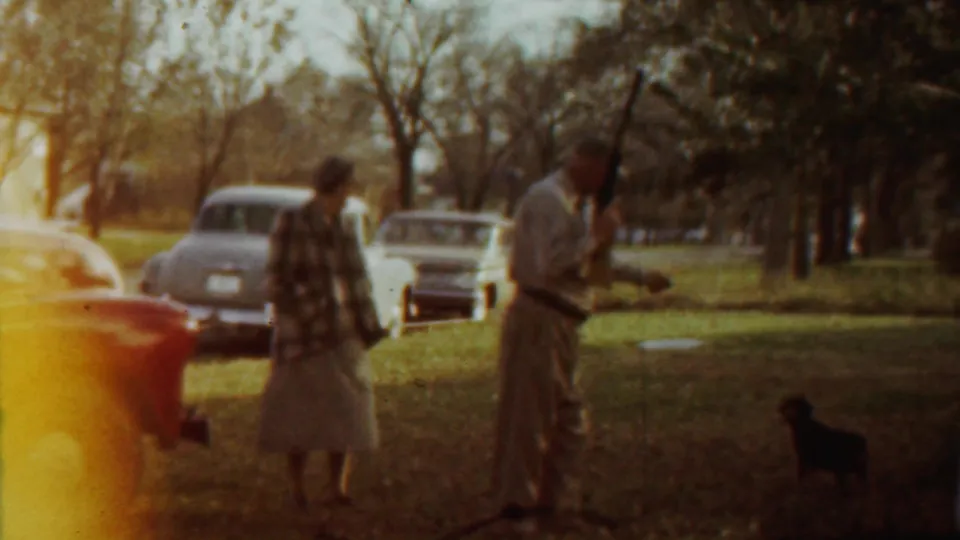 The wider park view shows the woman, uniformed man, small dark dog, and several parked cars in Saint Francis, Kansas.