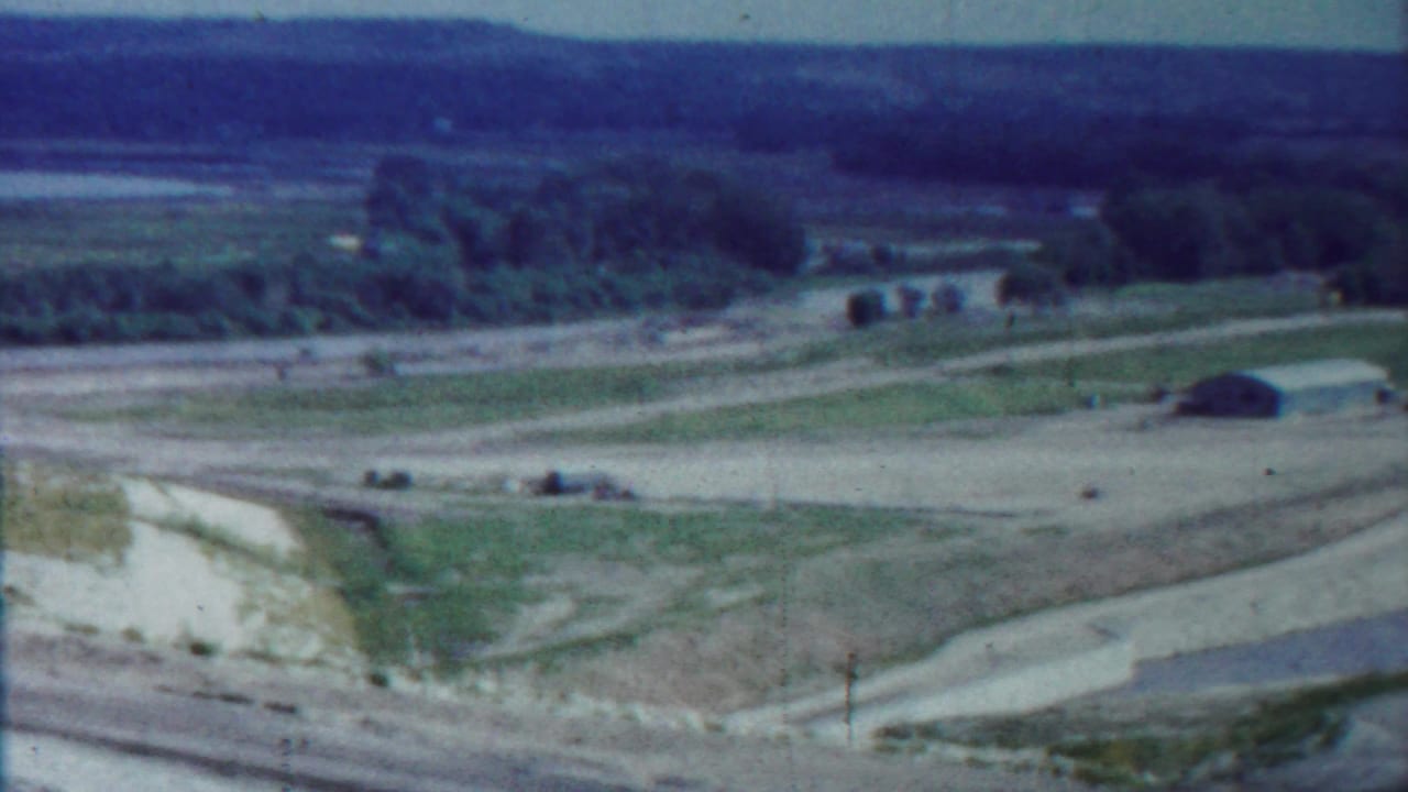 Wide rural landscape with dirt roads, green fields, trees, and distant water.